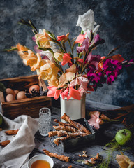 cookies in metal box with green apples, honey in small plate, crushed cookie on wooden board, eggs in wooden box, assorted gladiolus flowers in white vase and textile on blue background