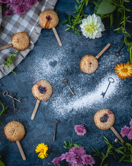 mini pies with berry filling on wooden sticks among flowers, old keys, textile with sugar powder over blue background, top view
