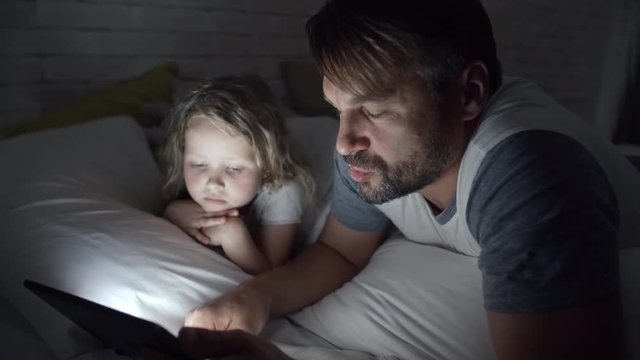 Tilt up of bearded father lying on bed with adorable little girl and reading her story on tablet in darkness