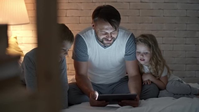 PAN of bearded father in nightwear sitting on bed with little boy and girl and reading digital book on tablet