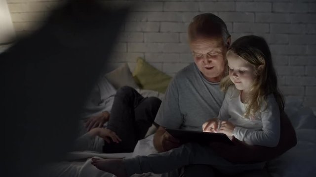 PAN of elderly woman in glasses lying on bed and chatting with grandson while cheerful senior man holding tablet and reading digital book to adorable little girl sitting on his lap