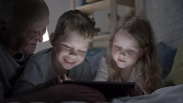 PAN of curious little girl lying on bed in bedroom with dim light and playing on tablet with laughing senior man adorable boy, then getting upset and turning away with crossed hands