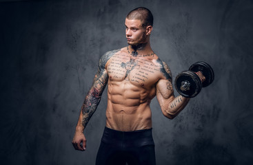 Shirtless, tattooed male holds dumbbell over grey artistic background.