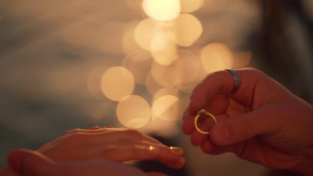 slomo extreme closeup hands of loving couple, man wearing engagement ring to his girlfriend at sunrise time with shining sea on background