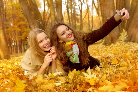 Two girlfriends are making a selphi on a pile of leaves in an autumn park.