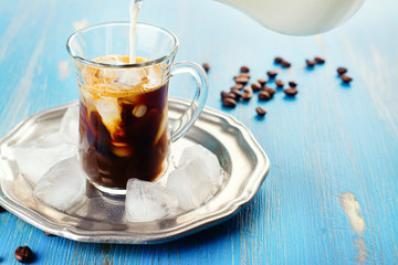Refreshing iced coffee with cream over blue summer background. Pouring milk into glass with coffee drink