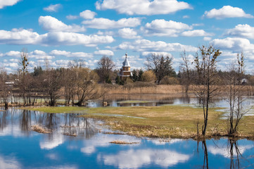 Fototapeta na wymiar Spring high water flooded agricultural fields and trees due to flood of river against background of the village and of Orthodox Church with reflection clouds and blue sky in water in temperate zone