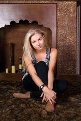 Pretty young woman sits on fur carpet in studio with decorative fireplace