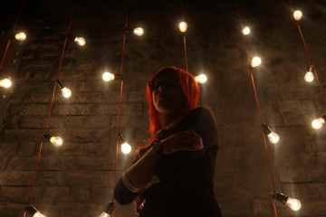 Fototapeta premium Girl in glasses poses near wall in black studio with garlands of lamps, under view