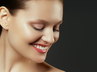 Fototapeta premium Pretty girl with big eyes and dark eyebrows smiling, a model with light nude make-up, gray studio background, beauty photo, close up. Beautiful spa model girl with perfect fresh clean skin