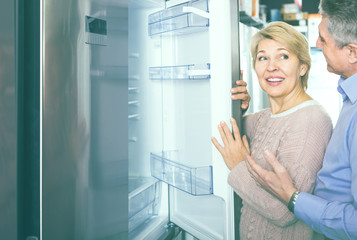 Fototapeta na wymiar positive mature married couple in shop of household appliances are choosing home modern refrigerator