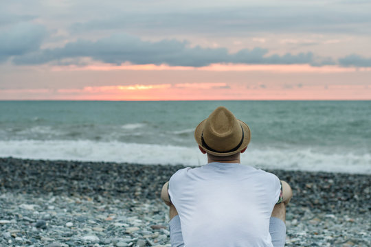 Young Man In Hat Sitting On The Beach Against The Backdrop Of The Sea And Sunset Sky. Back View