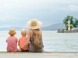 Mom and two sons sit on the pier and admire the sea and the mountains in the distance. Back view