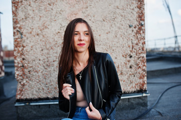 Portrait of a beautiful young woman in black leather jacket, jeans and sneakers sitting on a rooftop.