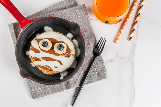 Funny food for Halloween. Kids breakfast pancake decorated like mummy with white chocolate sauce, banana, berries, with pumpkin smoothie juice, white table copy space top view