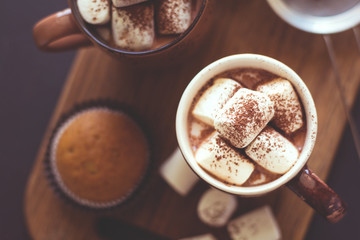 Cups with hot chocolate with marshmallow and muffins on dark wooden desk. Selective focus. Toned...