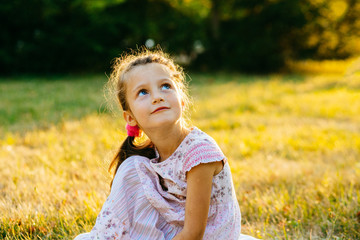 Cute preschooler girl with blue eyes sitting on the grass in sunset in summer time.