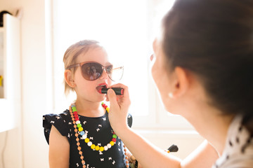 Mother painting lips of her cute little daughter with lipstick.