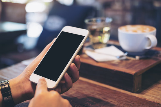 Mockup image of a woman's hand holding and pressing on white mobile phone with blank black screen with coffee cup on wooden table in vintage cafe