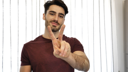 Attractive young man doing peace or victory sign with two fingers, indoor at home in his living-room