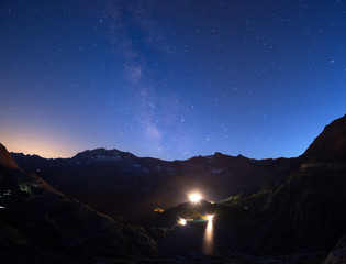 Obraz premium The colorful glowing Milky Way arch and the starry sky from high up on the Alps. Lights from hydroelectric lake dam.