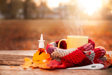 Tea cup covered with warm scarf, nose drops and thermometer on wooden surface - obrazy, fototapety, plakaty