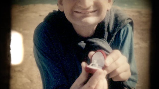 Fake 8mm amateur film: an ugly man kneels in front of her girlfriend at a romantic place and shows an engagement ring. Point-of-view of the woman. Funny scene.
