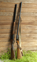 Two different (side-by-side & over/under) hunting double-barreled shotguns stand in the grass, leaning against the plank wall