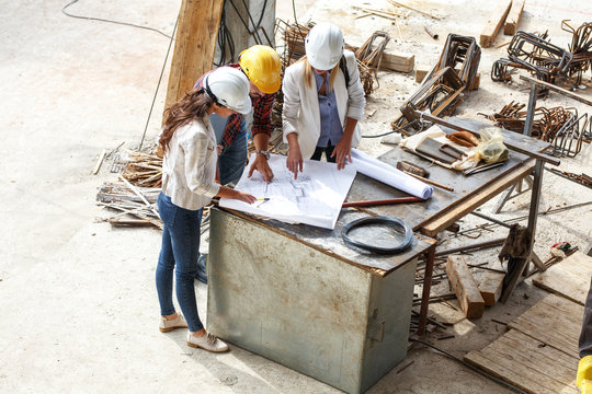 Two female  inspectors and architects discuss with head engineer about blueprints of construction site.