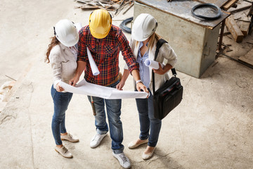 Two female  inspectors and architects discuss with head engineer about blueprints of construction site.