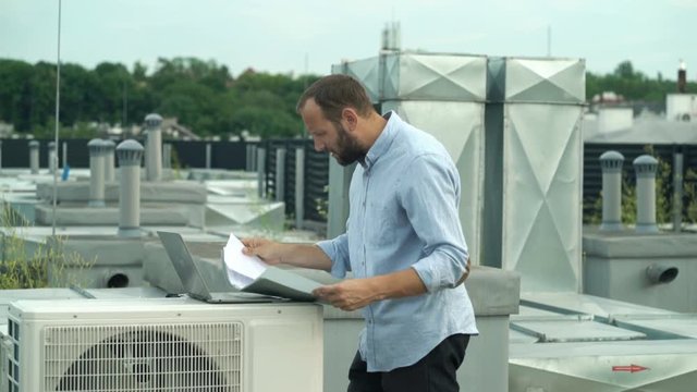 Upset, unhappy man with laptop and documents checking air conditioning plan on roof
