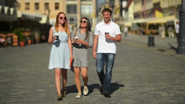 Three Happy Friends Drinking Coffee while Walking around the City. Full Height Portrait of Two Girls and One Boy with Beverages in the Hands.