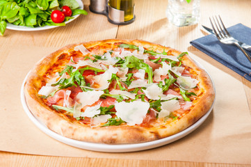 Pizza with prosciutto, parma ham, arugula and parmesan on served wooden restaurant table. Italian...