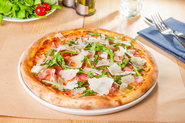 Pizza with prosciutto, parma ham, arugula and parmesan on served wooden restaurant table. Italian cuisine. Selective focus