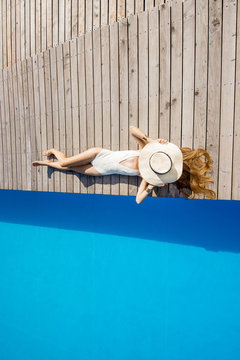 Woman lying with sunhat on the poolside near the basin with blue water. Geometric top view with copy space