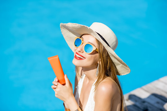 Portrait of a young woman in sunhat and swimsuit holding a tube with sunscreen lotion at the swimming pool