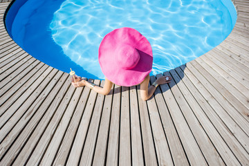 Woman in big pink sunhat with cocktail drink relaxing at the round swimming pool with blue water...