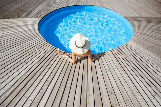 Woman in big sunhat with cocktail drink relaxing at the round swimming pool with blue water outdoors. Top view