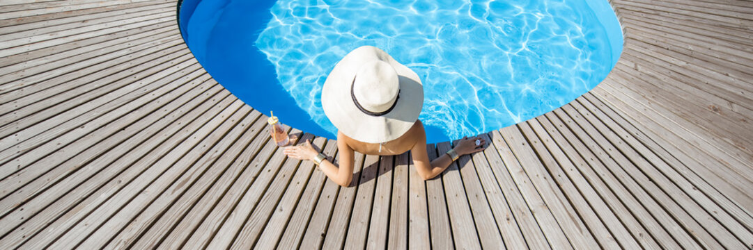 Woman in big sunhat with cocktail drink relaxing at the round swimming pool with blue water outdoors. Top view