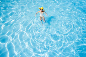 Young woman in yellow sunhat swimming at the basin with blue water. General view from above