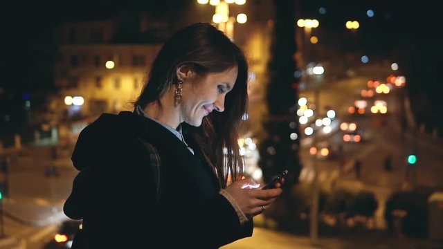 Young beautiful woman standing in the city centre on the background of traffic road in the evening and using smartphone.