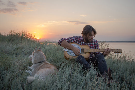 Young handsome men with blues guitar with husky dog having good time, sunset in lake background 