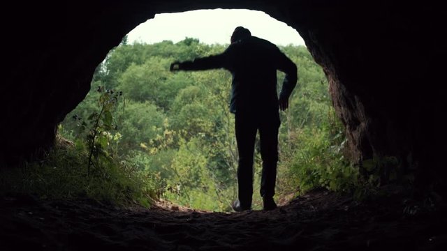 Traveler exits from dark cave chamber from darkness to day light, stops and raises his hands up. A look from the cave. Deep forest at background. Freedom travel adventure theme