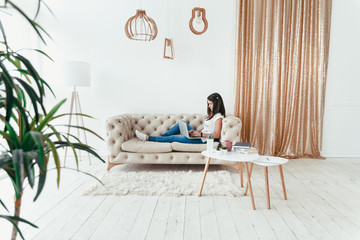 Beautiful young woman working from home on a laptop lying on the sofa in the room