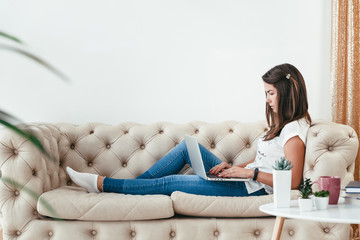 Beautiful young woman working from home on a laptop lying on the sofa in the room