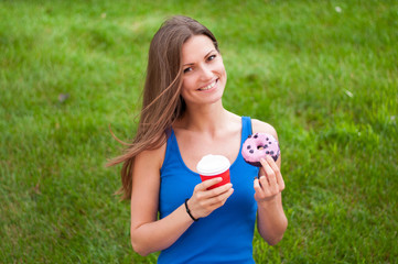 Smiling young woman eating a donut and holding a Cup of coffee outdoors

