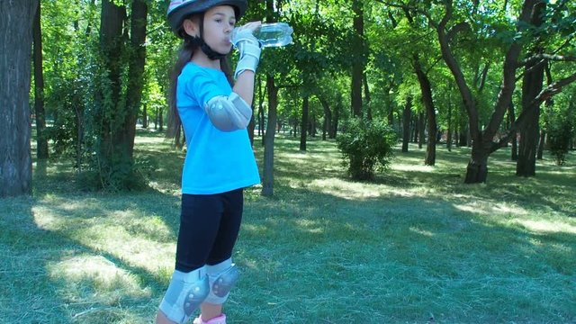 A little girl on rollers drinks water. A child in protection and helmet drinks water in the park.