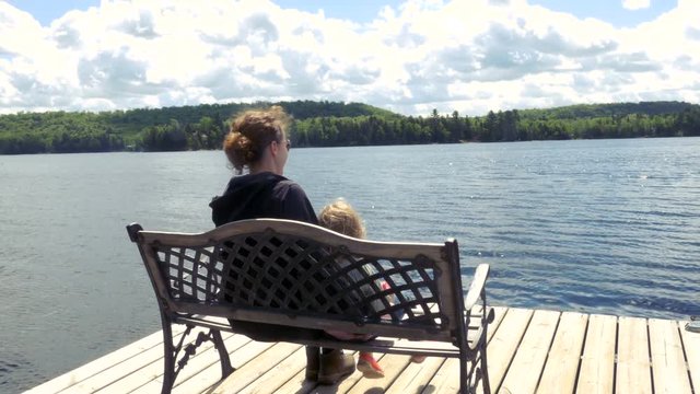 A mother and daughter sitting on a bench atop a pier in Georgian Bay, Ontario. Medium shot.