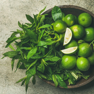 Flatlay of freshly picked organic limes and mint leaves for making cocktail or lemonade in wooden plate over grey concrete stone background, top view, square crop