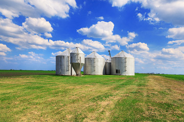 Fototapeta na wymiar Metal grain bins on farmland in a rural background. Blue and clouds in the background.